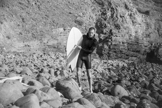 Andrea in a wetsuit holding a fish surfboard on rocky terrain